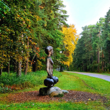 Attēlā ir koka skulptūra joga, cilvēks sēž jogas pozā, apkārt koki