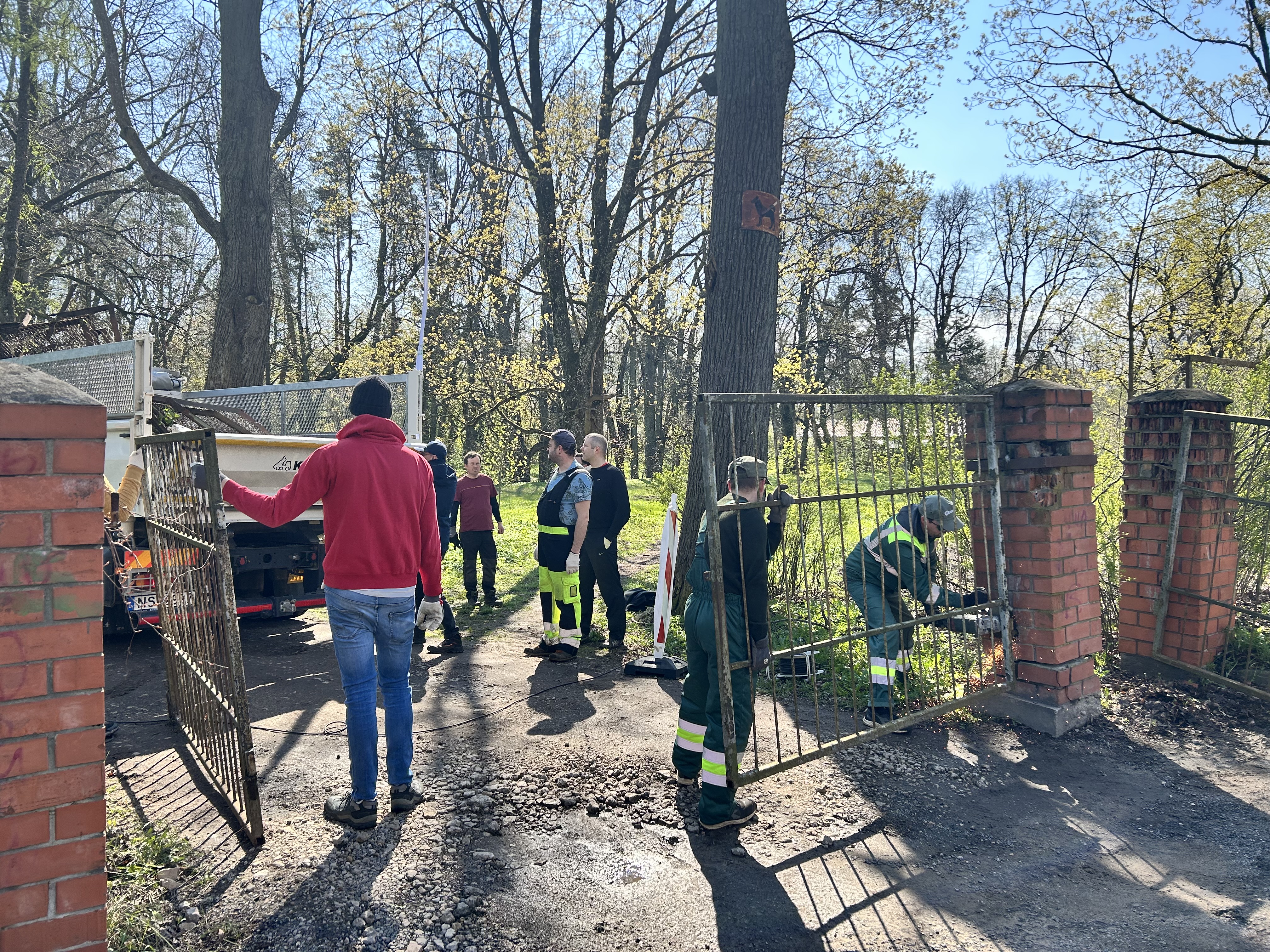 Cilvēku grupa demontē vecos vārtus, klāt stāv lielā mašīna, kurā tos pēc tam ieraut