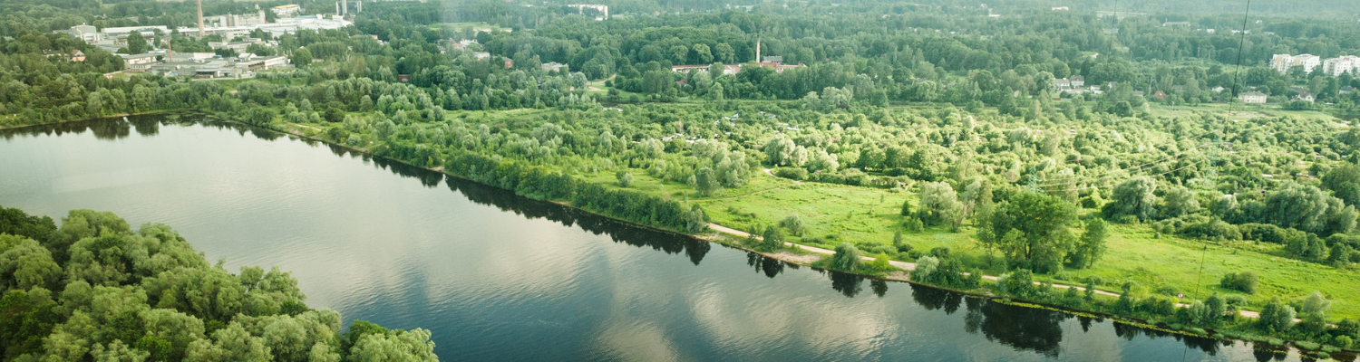 Attēls uzņemts ar dronu, tā vidū ir upes līcis, tam apkārt ir zaļojuši koki, tālumā redzama rūpnieciska apbūve